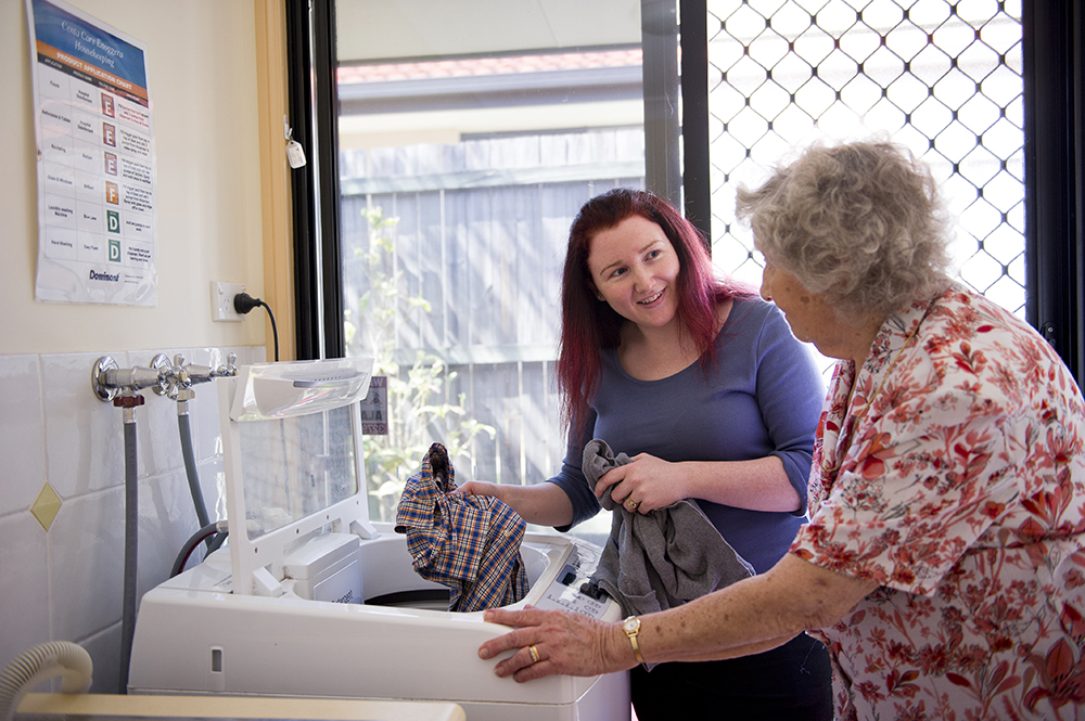 A Woman Helps An Old Woman Put Clothes In The Washing Machine - Home Care Solutions - ESP Healthcare